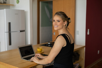 Young woman working at home in her kitchen with laptop and papers on kitchen wooden desk. Home office concept.