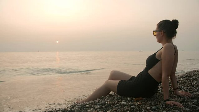 A woman in sunglasses relaxes on a pebble beach at sunset.