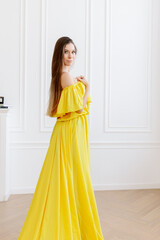 Portrait of a pretty and attractive brunette European girl in a bright yellow silk dress. The model is standing with her back to the long train of the dress with waves on the back.