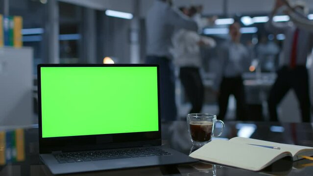 Focus on laptop with green screen. Colleagues dance and have fun on blurred background
