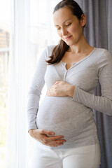 caucasian happy pregnant woman with hands on belly standing near window