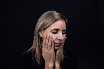 Depressed woman face with hands touching cheeks wearing black dress on black background. Victim of physical and psychological abuse. Gaslighting. Relative aggression.