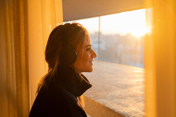 Side view of young woman listening to music with headphones in her ears by the window with a city view landscape in the background. High quality photo