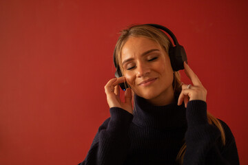 Young woman listening to music with headphones in her ears in his apartment. Red painted wall in the background. High quality photo. Mobile phone, technology, music, urban living, feel good concept.