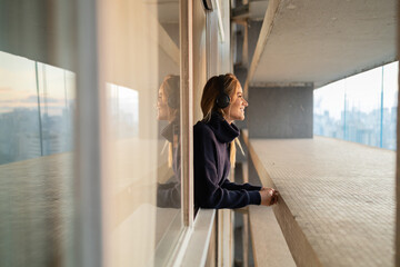 Side view of young woman listening to music with headphones in her ears by the window with a city view landscape in the background. High quality photo