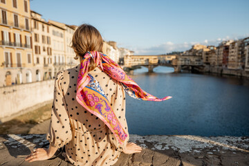 Young woman enjoys beautiful view on famous Old bridge in Florence, sitting back on the riverside at sunset. Female traveler visiting italian landmarks. Stylish woman wearing dress and colorful shawl
