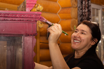 Blissful woman engaged in painting wooden cupboard with fantastic ornaments in pink with paint...