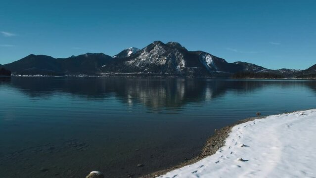 Scenic Bavarian lake Walchensee in south Germany in the alps mountains close to Austria. Blue clear drinking water waves at the white shore and idyllic blue sky in winter. 4K UHD drone aerial.