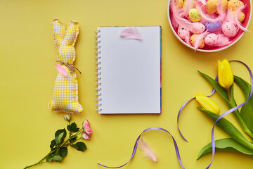 Festive Easter mock up notepad. Pink bowl with colorful eggs, pink feathers, handmade cute bunny and flowers. Happy Easter concept. Post card mock up on yellow background
