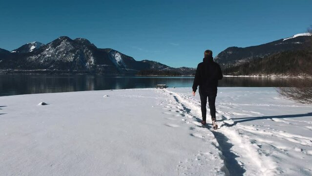 Young man on a beach covered in snow at Lake Walchensee in Bavaria, south Germany in the alps mountains close to Austria. Blue clear drinking water waves at the white shore and blue sky in winter.