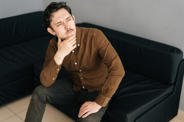 Portrait of pensive young man with little moustache holding hand on chin and thoughtful looking up, thinking over solution, sitting in sofa on background of grey wall.