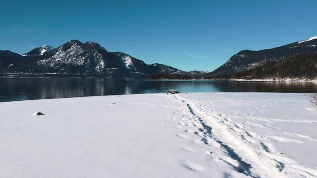 A beach covered in snow at Lake Walchensee in Bavaria, south Germany in the alps mountains close to Austria. Blue clear drinking water waves at the white shore and blue sky in winter. 4K drone aerial