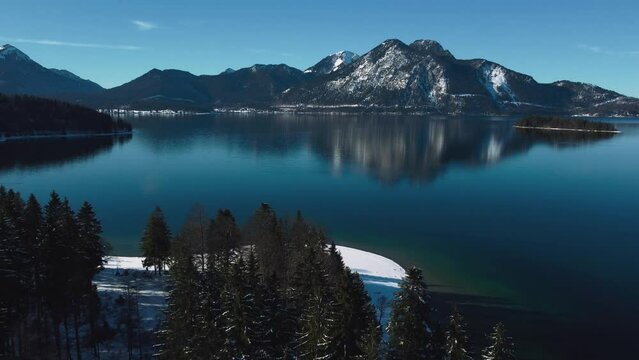 Famous Bavarian lake Walchensee in south Germany in the alps mountains close to Austria. Blue clear drinking water waves at the white shore and idyllic blue sky in winter. 4K UHD drone aerial.