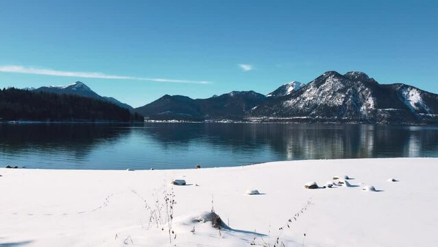 Beach covered in white snow at Lake Walchensee in Bavaria, south Germany in the alps mountains close to Austria. Blue clear drinking water waves at the shore and blue sky in winter. 4K drone aerial