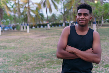 A young African man in sportswear, standing in a park, smiling with his arms crossed.