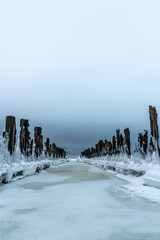 Old wooden pier at the Kupskalnu nature trail in Latvia by the Baltic Sea during cold winter morning. Frozen pier, ice, snow, moody clouds