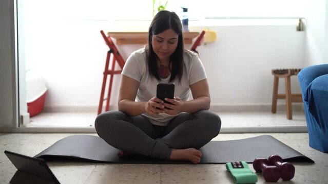 Chica joven guapa con mallas grises mirando una tableta y el smartphone mientras hace ejercicio en casa