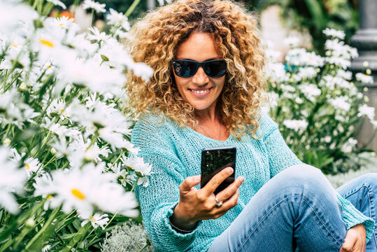 Cute Caucasian Woman Using Mobile Phone Outdoor Sitting With Daisies Spring Flowers Around. Happy Female People Chat And Send Message On Celular Device In Leisure Connection Activity