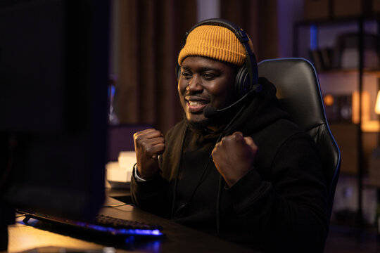 Dark room lit by led lights man sitting in front of computer wearing sweatshirt and yellow cap headset. Music producer's studio rapper processing tracks satisfied with his work clenches fist with joy - Powered by Adobe