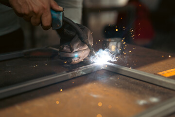 A male worker is welding electricity to a steel workpiece. causing sparks and white smoke