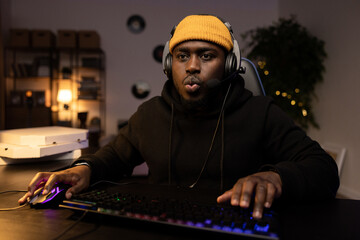 A focused guy in a yellow cap with a headset is playing a video shooter carefully on a computer. Online multiplayer championship. In the background a dark room at night with a mess of pizza boxes