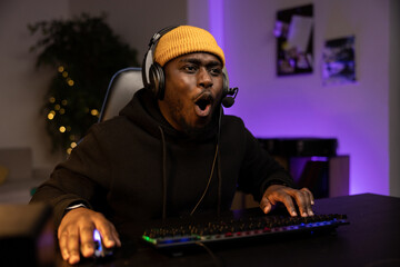 Shocked surprised male professional computer gamer opens mouth while looking at computer screen talks to team through headset presses buttons of backlit keyboard. Dark room lit by purple led lights