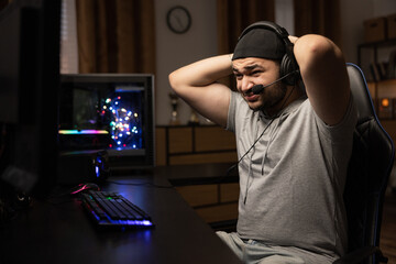 A man playing professionally on a computer with a headset puts his hands behind his head in anger, disappointment, lost a round in a video game, a middle-aged guy in a dark room © ABCreative