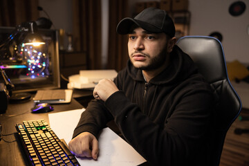 Shocked puzzled programmer IT specialist hacker internet criminal in black sweatshirt and helmet is terrified looks into the computer monitor, in the background dark room