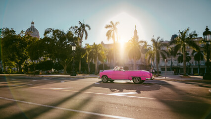 Havana, Cuba. Różowy cabriolet w świetle zachodzącego słońca.  Jedzie ulicą miasta pod palmami i niebieskim niebem