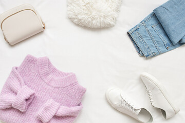 Lilac knitted sweater, blue jeans, white sneakers, bag and fur scarf lie on white background. Overhead view of woman's casual outfit. Trendy stylish women clothes. Flat lay, top view.