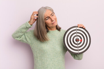 middle age woman feeling puzzled and confused, scratching head. darts target concept