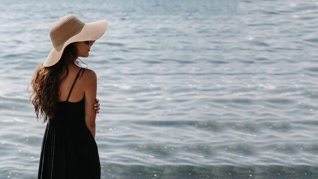 A picture of a beautiful young woman with long dark hair in a dress and a straw hat, posing on the beach, standing with her back and looking at the endless sea. A place for your text.