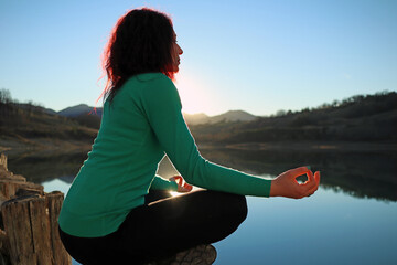 young caucasian girl dressed in green meditates above a log on the lake at sunset
