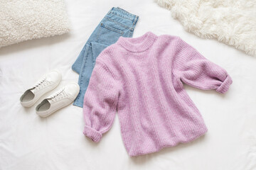 Lilac knitted sweater, blue jeans and white sneakers lie on white background. Overhead view of...