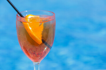 Glass with orange cocktail and orange slice against the blue water of the pool