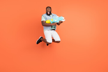 Photo of excited afro guy wear denim vest shooting water gun jumping high isolated orange color background