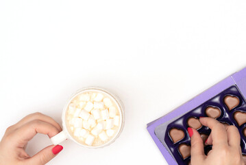 Female hands with a cup of cappuccino with white small marshmallows and a box of heart shaped chocolates on white background. Valentines day, love concept. Flat lay style with copy space