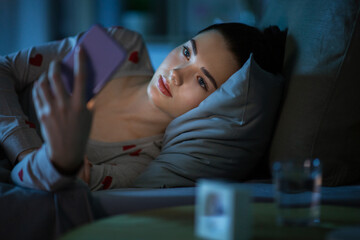 technology, bedtime and rest concept - teenage girl in pajamas with smartphone lying in bed at night