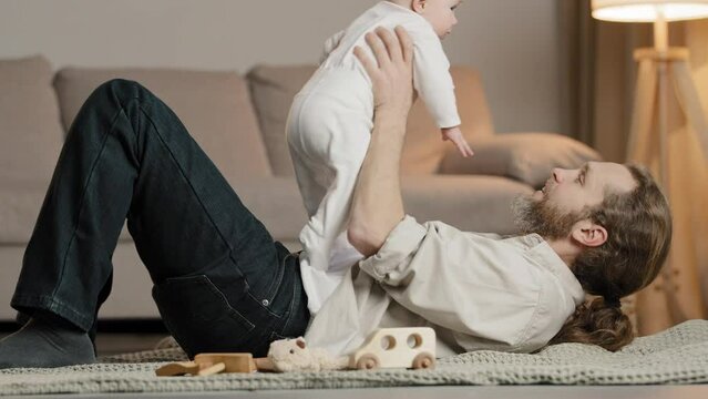 Caucasian family at home adult bearded father dad lying on floor with little daughter son baby playing with child lifting infant up talking with newborn spending time together care fatherhood concept