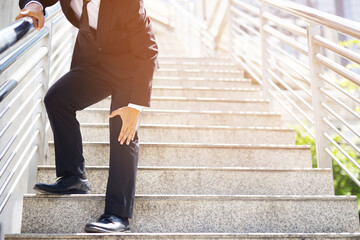 Businessman in a suit have the sore pain and painful knee problem expression and walk up and down on the stairway during go to office. Health care concept.