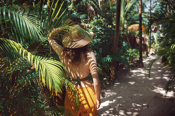 Young beautiful woman stands in the shade of palm trees, view from the back. A girl walks among tropical greenery on a bright sunny day in Mexico, Tulum - obrazy, fototapety, plakaty