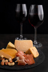 Blurred image in the foreground of different cheeses, jamon, in the background glasses with wine on a black background.