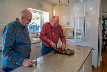 An older LGBTQ+ man is lifting freshly baked muffins out of the pan to let them cool as his husband watches.