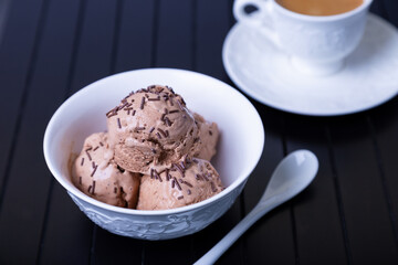 Chocolate ice cream with chocolate sprinkle in a white bowl on a black background. Three balls of homemade ice cream and a cup of coffee. Close-up.
