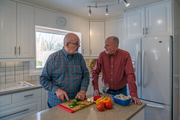 A mature LGBTQ+ couple engage in conversation in their kitchen as one of them slices a cucumber and prepares food.  The other is watching and drinking wine.