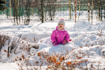 Little caucasian girl in bright pink warm clothing playing smiling sitting in snow in sunny winter day. Horizontal shot, front view. Copy space. Happy childhood and winter leisure activity concept.