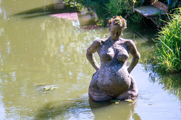 Sculpture of a woman with a big belly and plump legs nude made of stone stands in the water.