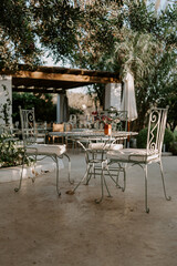 A rustic outdoor dining setup in Ibiza, featuring a shaded seating area with white wooden furniture under a canopy. Surrounded by olive trees, this cozy spot blends nature and Mediterranean charm