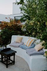 A cozy Mediterranean outdoor lounge with a built-in white bench, soft cushions, and lush greenery. The inviting space exudes warmth, relaxation, and rustic charm, perfect for slow living and leisure