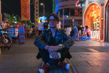 Fototapeta premium Portrait of young stylish brunet caucasian man in black leather jacket and yellow sunglasses Sitting on the street at night. High quality photo
